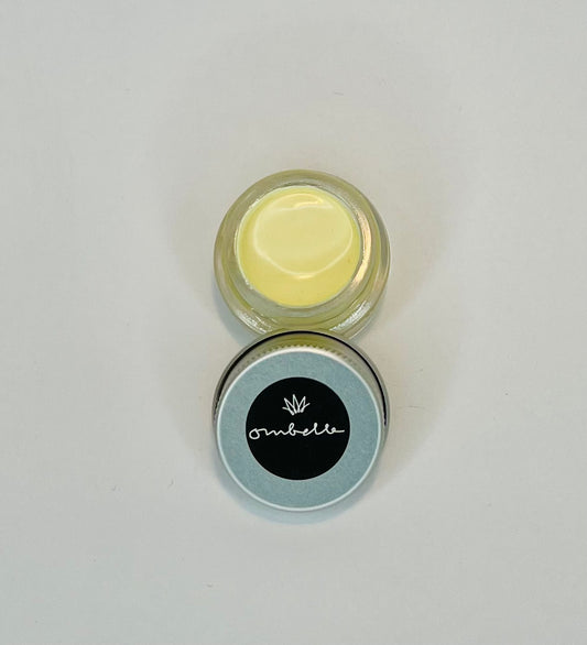 Glastiegel geöffnet mit Gesichtscreme mit LSF 30. Der Deckel ist am Tiegel gelehnt und man kann das Ombelle Logo mit weißer Krone und schwarzem Hintergrund sehen. Die Farbe der Creme ist hellgelb.