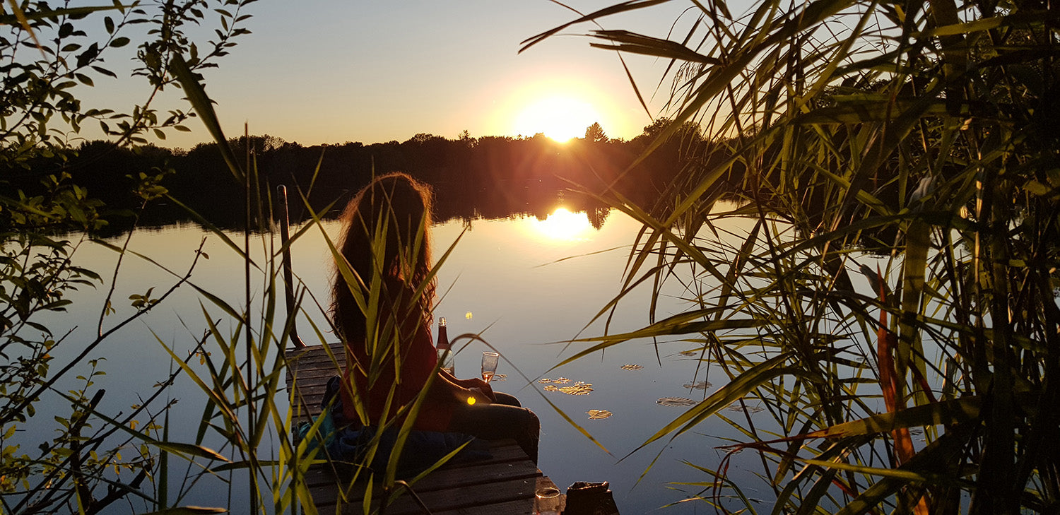 Ombelle luxuriöse Naturkosmetik, Gründerin Cécile Morice auf einem Steg am See bei Sonnenuntergang. Mit Champagner Glas und Wasserblumen, Bambus im Vordergrund. Frau mit langem Haaren in der Dämmerung