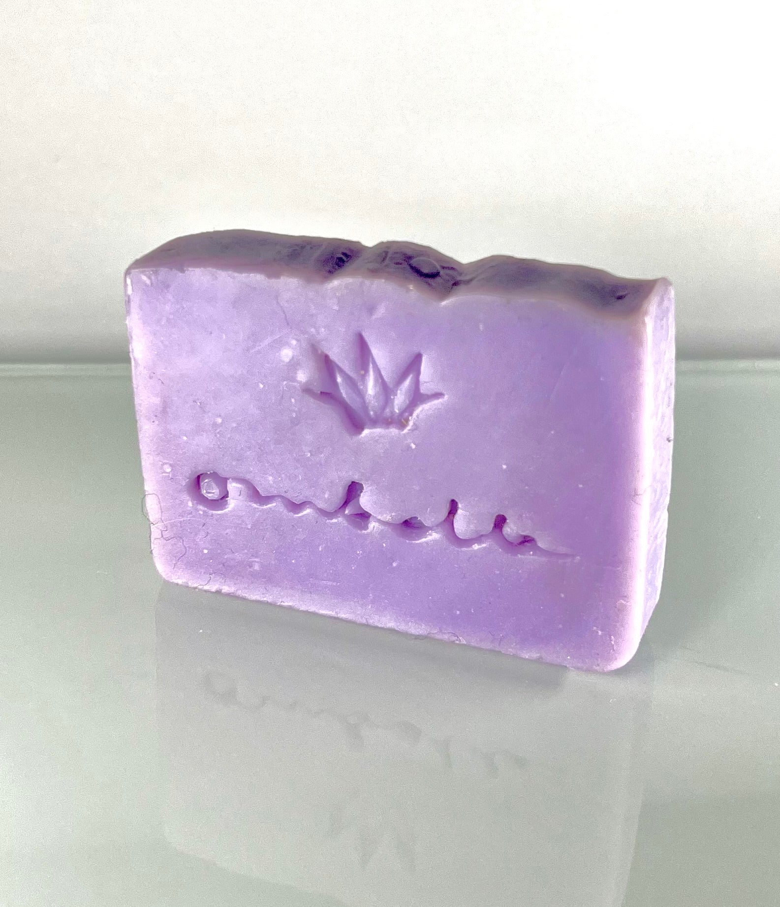Vegane Naturseife Lavendel fotografiert auf eine Glasplatte, die Seife spiegelt sich im Glas wieder. Die Farbe ist Lila und die Seife ist mit dem Ombelle Logo geprägt.