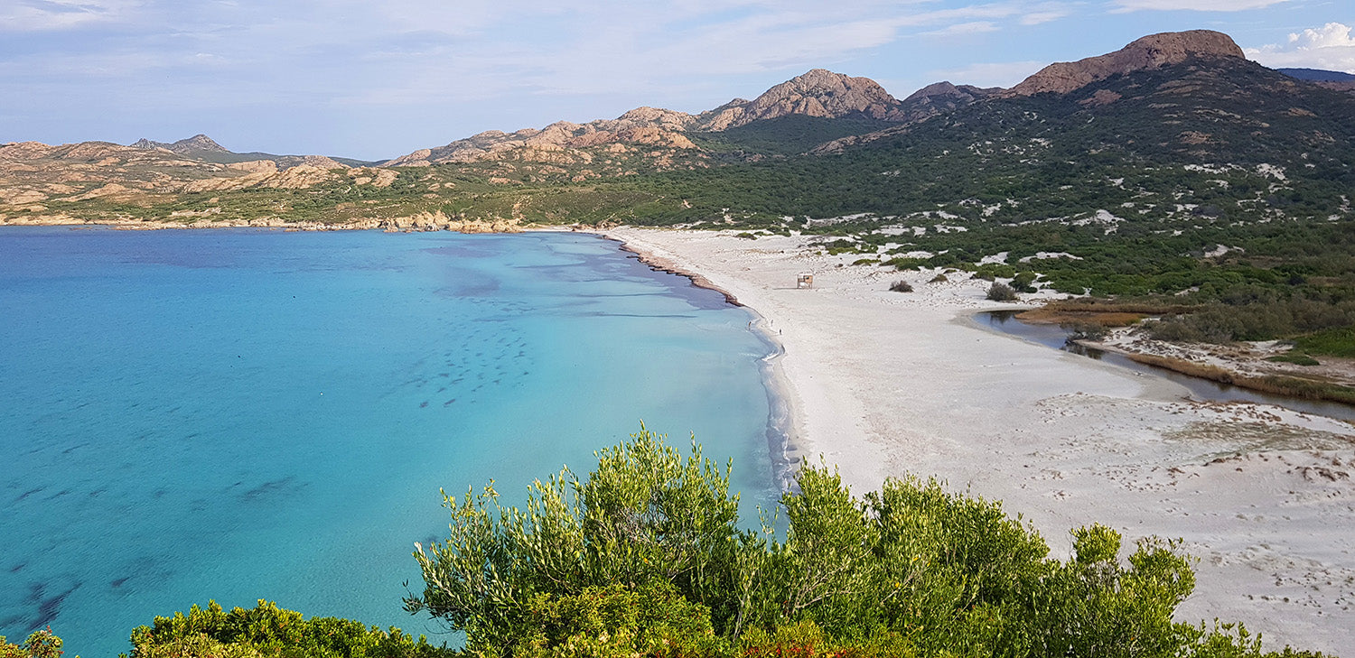 Ombelle Naturkosmetik Strandansicht an französischer Küste.Einsamer Strand mit weißem Sand und  mit Bergen im Hintergrund. Azurblaues Wasser. Ruhiges Meer an der Côte d'Azur der französische Riviera. Grüne Bäume im Vordergrund.