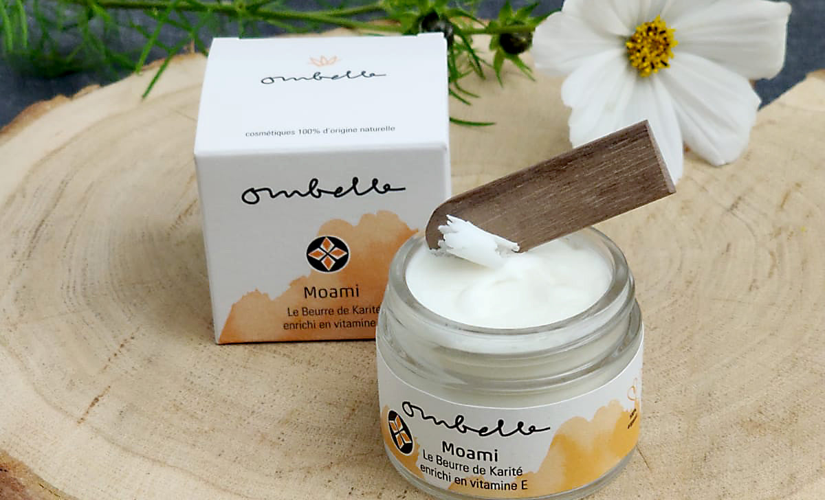 Ombelle Moami Sheabutter Glastigel geöffnet mit Holzspatel auf Holzscheibe mit weißer Blüte Fair Trade Bio 100% natürlich und bio zertifiziert La nouvelle Cosmétique