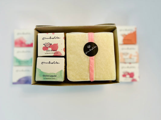Geschenkbox mit einer Seife mit rosaner Schleife und schwarzem runden Etikett. Im Hintengrund sind man jeweils drei kleine Deocremes und drei Lippenplege unscharf.