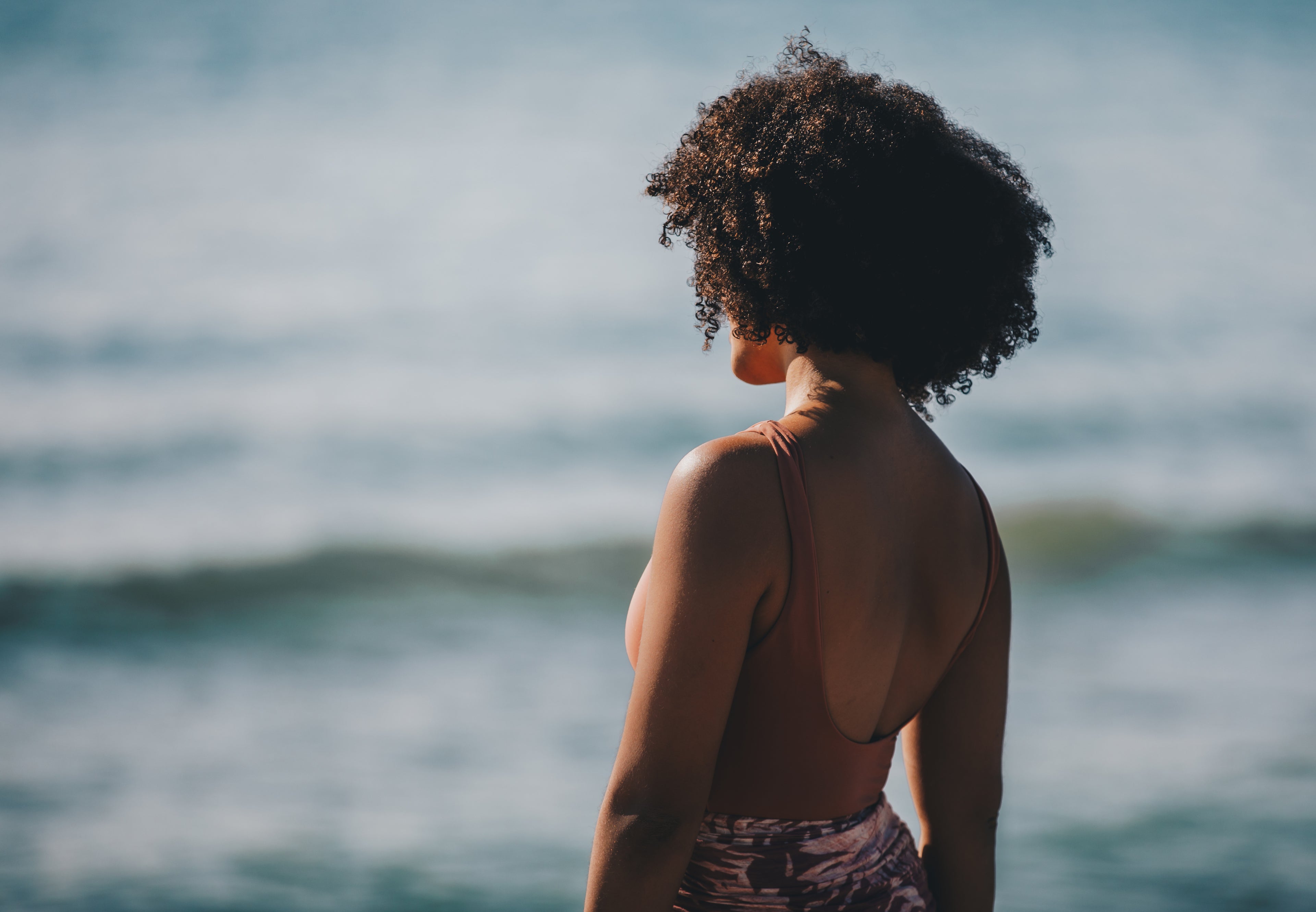 Ombelle Bio Fair Trade Shea Butter Frau mit lockigem Haar steht am Strand und blickt zum Meer hinaus. Rückenausschnitt mit Pareo. Dunkle Frau mit lockigem Haare. Blaues Meer mit Wellen und reflektiertem Sonnenscheine